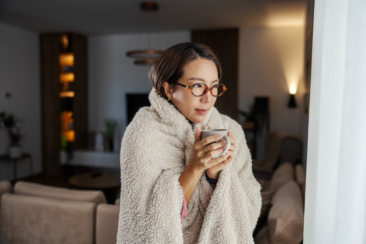 Woman wrapped in cozy blanket at home, sipping a hot mug and gazing out the window in a quiet, contemplative moment